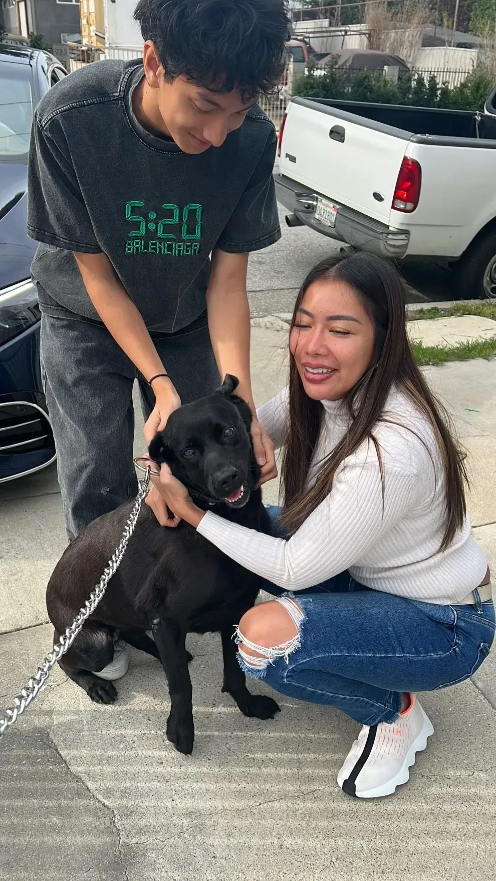 Two people crouch with a black dog on a sidewalk beside parked cars.