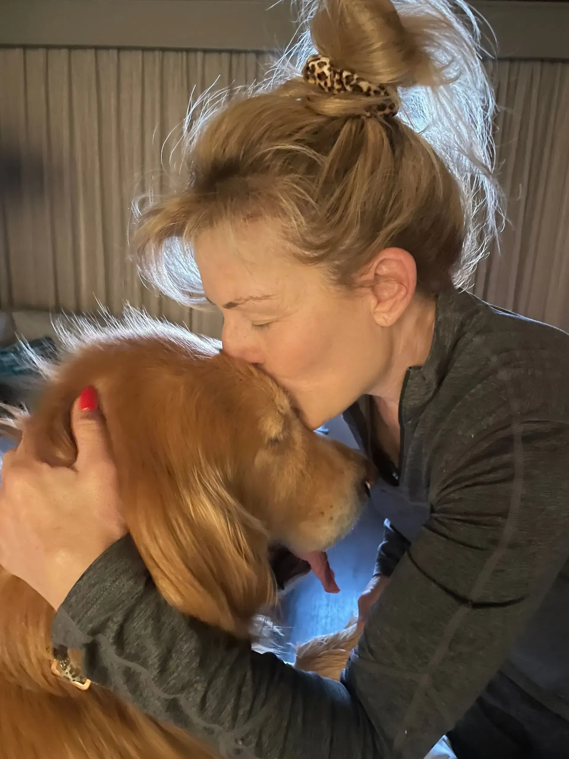 Woman gently hugging a golden-brown dog indoors by a window with curtains