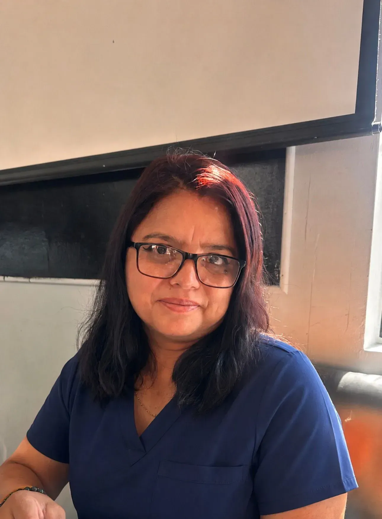 Woman in blue scrubs and glasses, seated indoors near a window, looking at the camera.