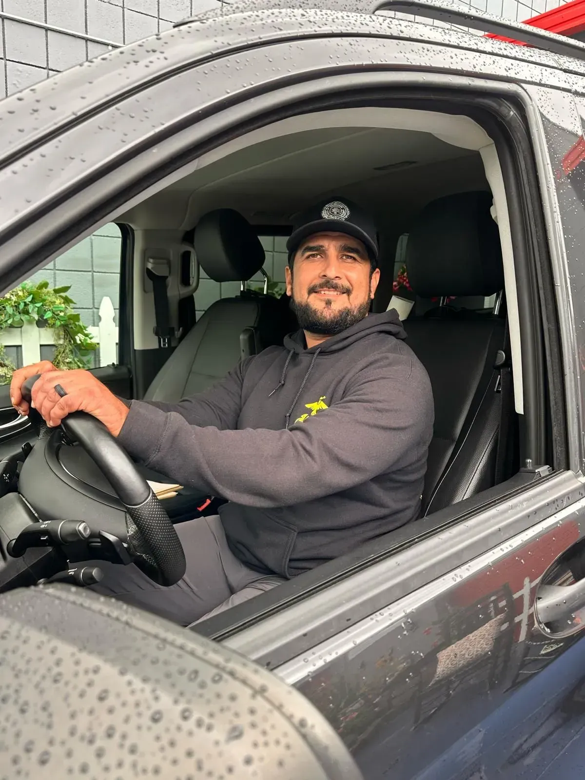Man smiling in a dark jacket and cap, seated in a car with rain drops on the window.
