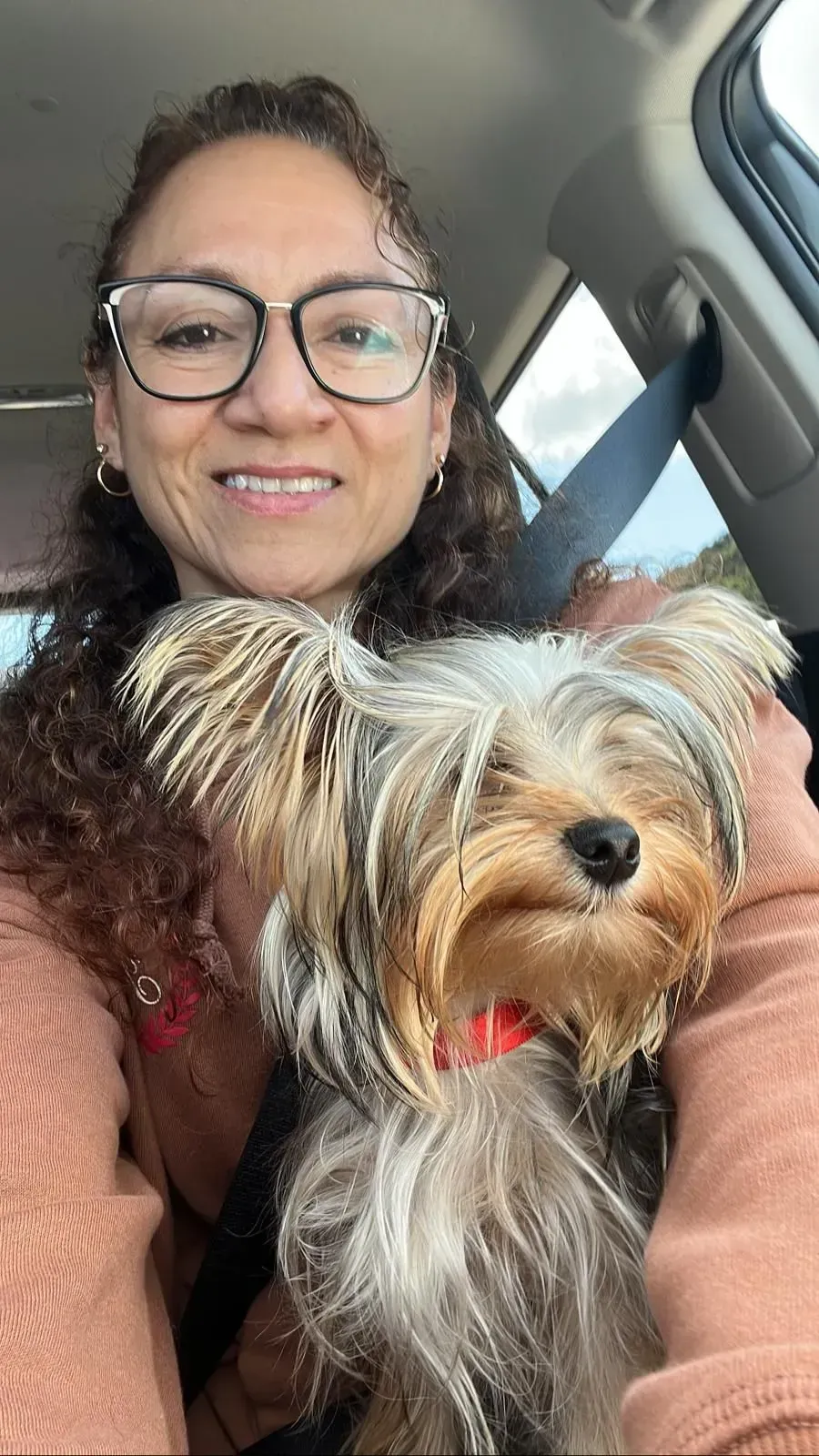 Woman smiling in a car, holding a small Yorkshire terrier with a red bow tie.