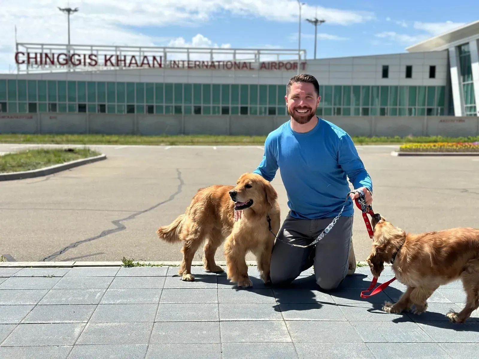 Man kneels with two golden dogs and a brown dog outside Chinggis Khaan International Airport.