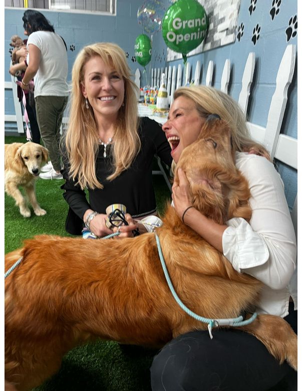 Two women smile and hug a large golden retriever at a dog event with a "Grand Opening" sign.