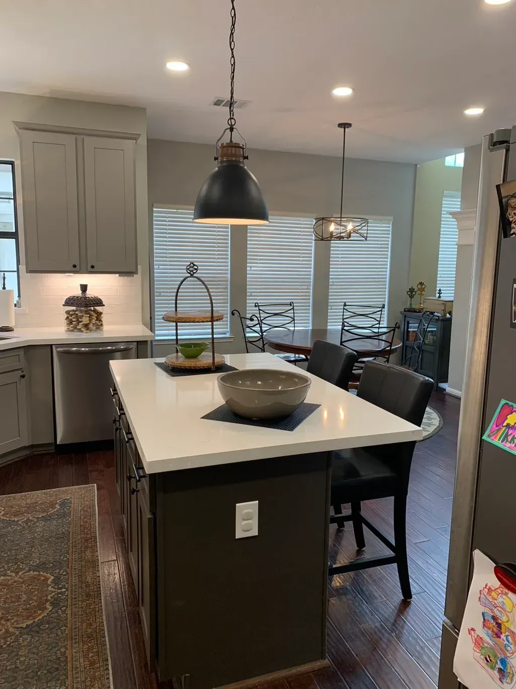 Modern kitchen with island, black pendant light, bar stools, and dining area by windows.