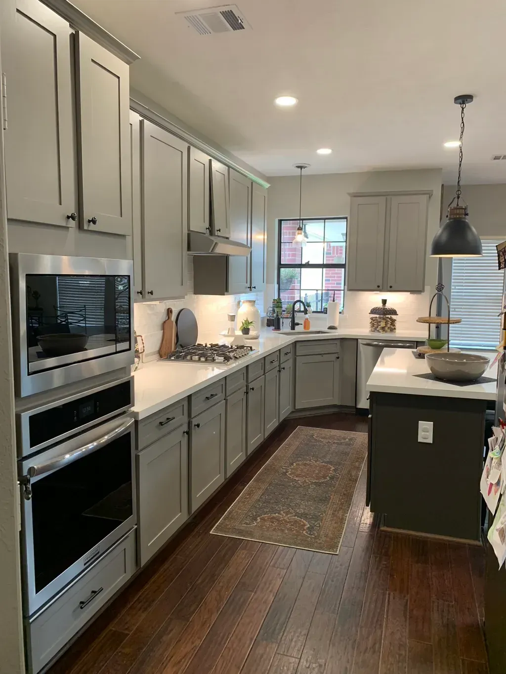 Bright modern kitchen with gray cabinets, wood floor, island, pendant light, and built-in ovens.
