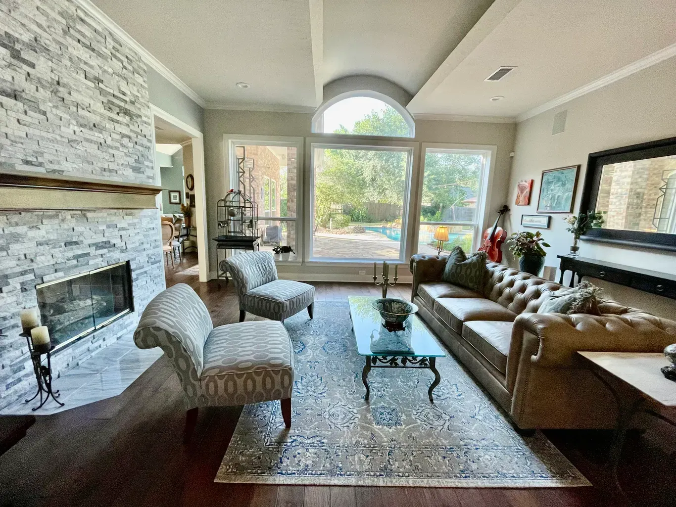 Bright living room with stone fireplace, tan sofa, chairs, and large sunlit windows overlooking greenery