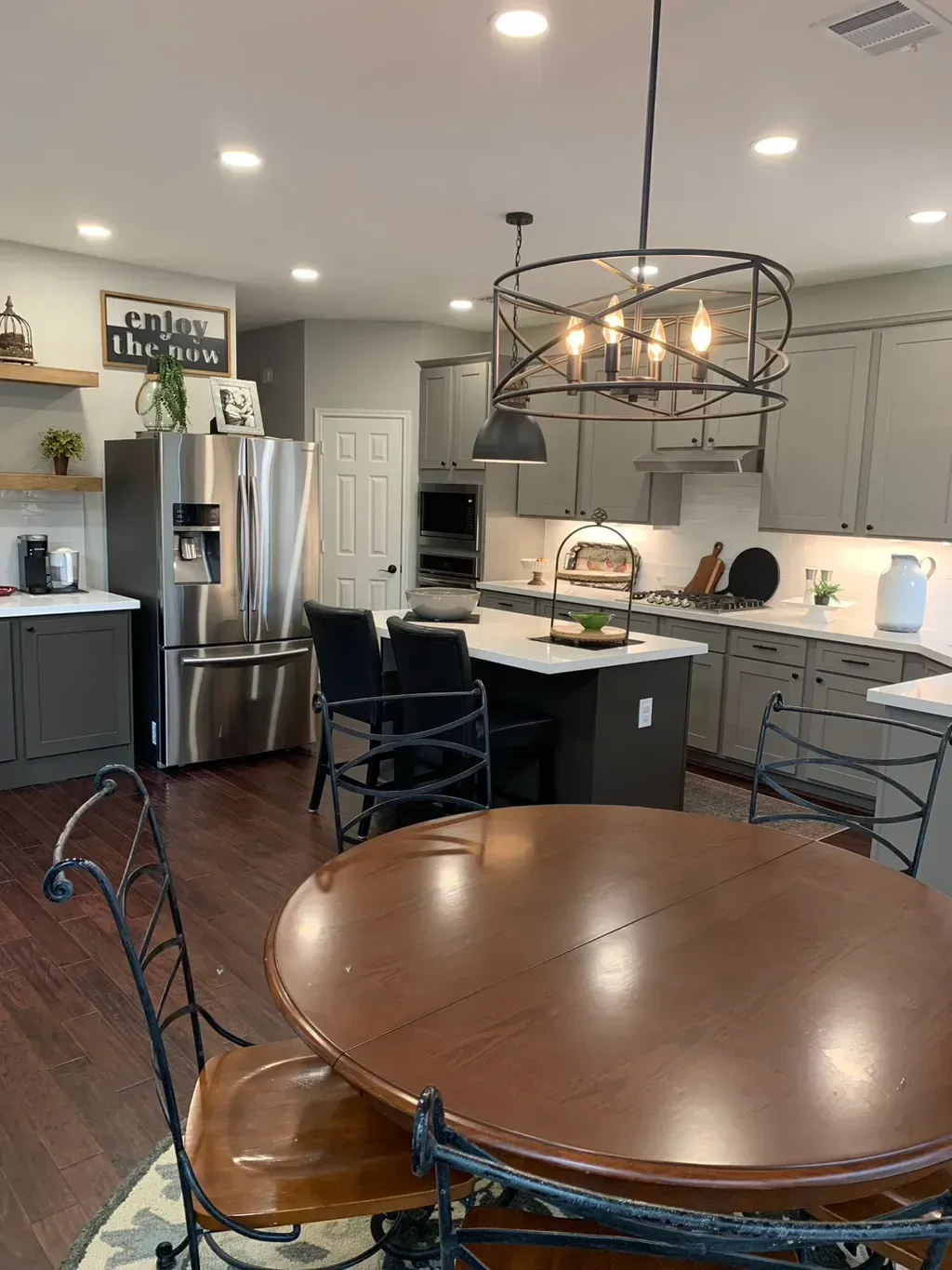 Bright kitchen with stainless fridge, white cabinets, round wooden table, and black chandelier.