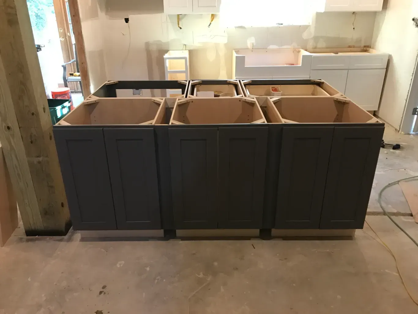 Dark kitchen island cabinets being installed in a room with unfinished countertops and a sink behind it