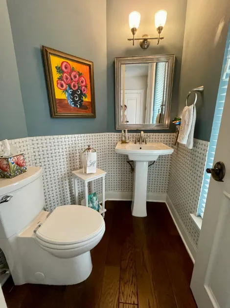 Small bathroom with white pedestal sink, toilet, dark wood floor, wall art, and a mirror above the sink.