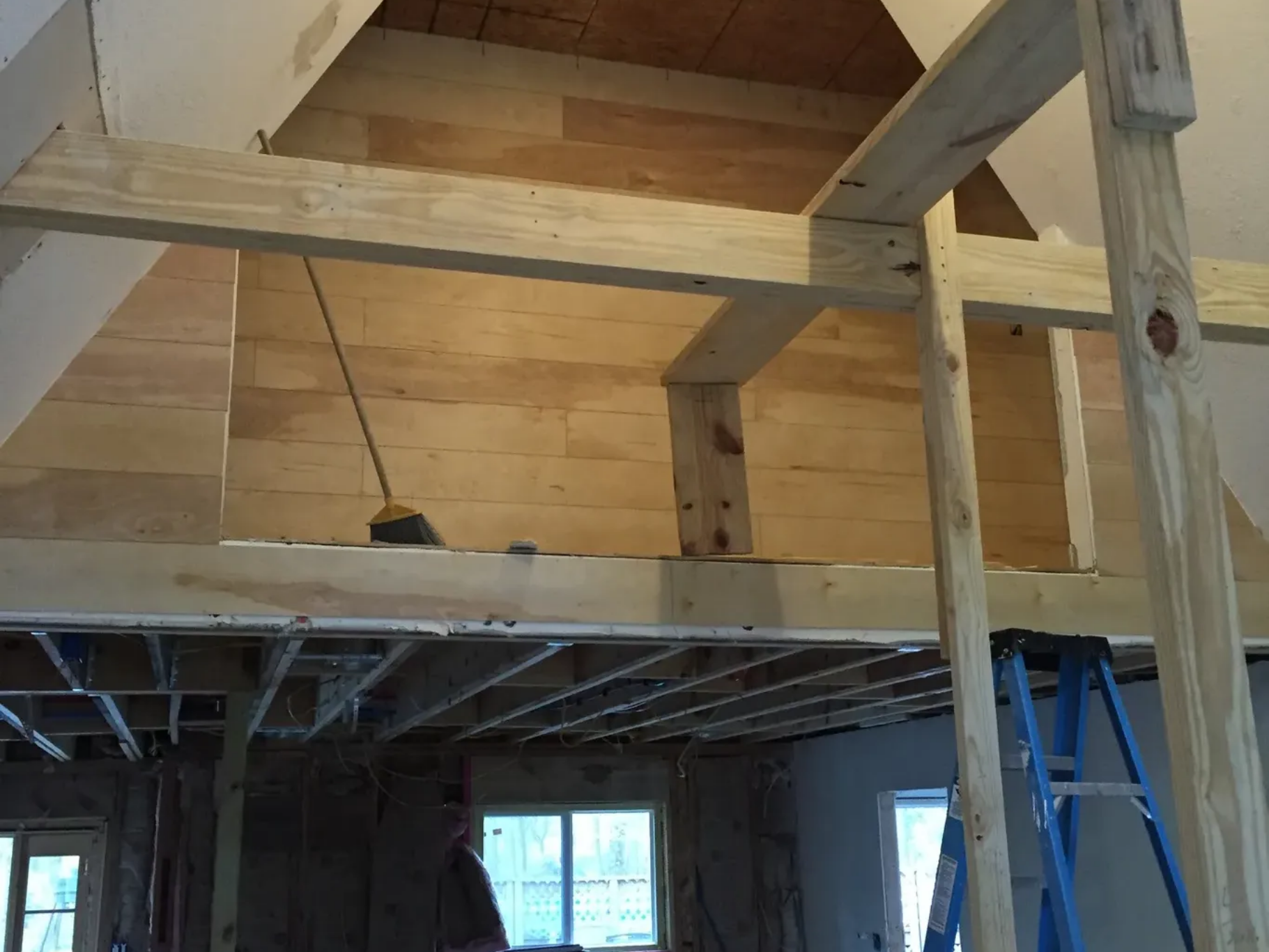 Interior under construction with exposed beams, wood framing, and a blue ladder in a room with windows.