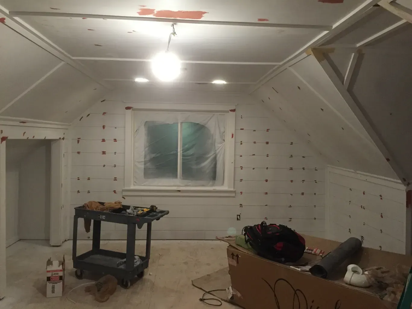 Unfinished attic room with white insulation panels, a lit ceiling bulb, window, and a work table with supplies.