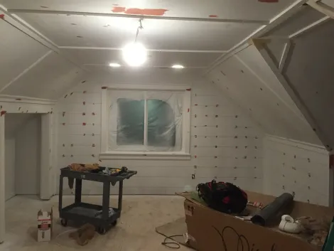 Unfinished attic room with white insulation panels, a lit ceiling bulb, window, and a work table with supplies.