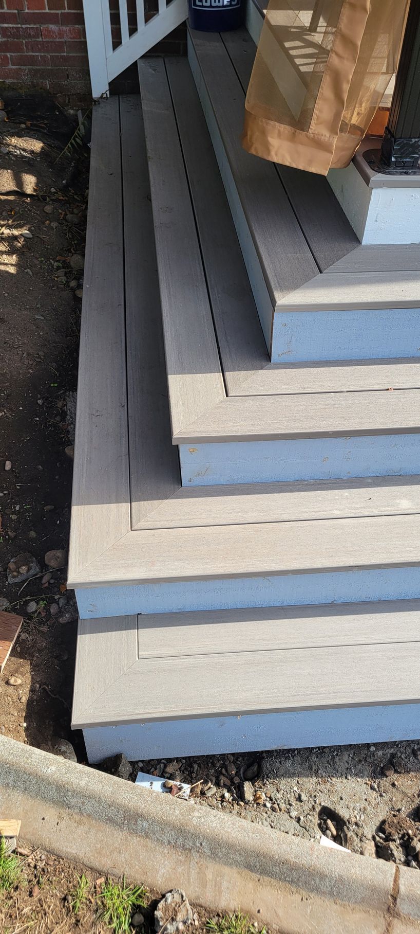 A side view of wooden steps with gray treads and white risers leading up to a porch, surrounded by soil and a stone border.