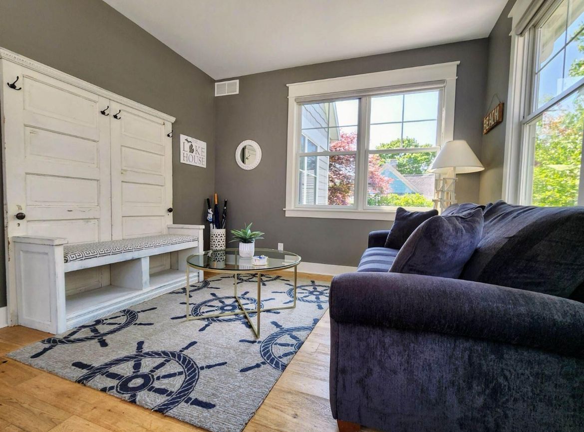 Bright living room with gray walls, blue sofa, white bench, patterned rug, and large sunlit windows
