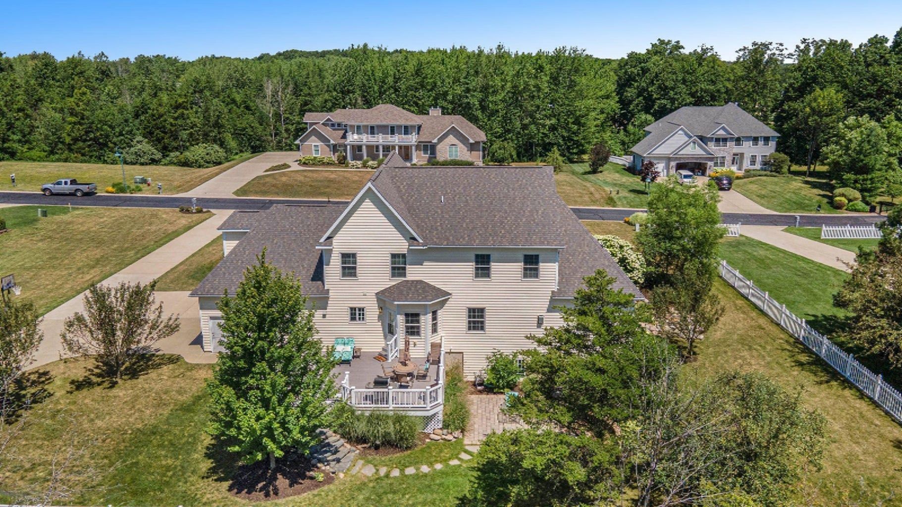 Aerial view of a beige house with a gray roof, surrounded by trees and neighboring homes in a suburban area