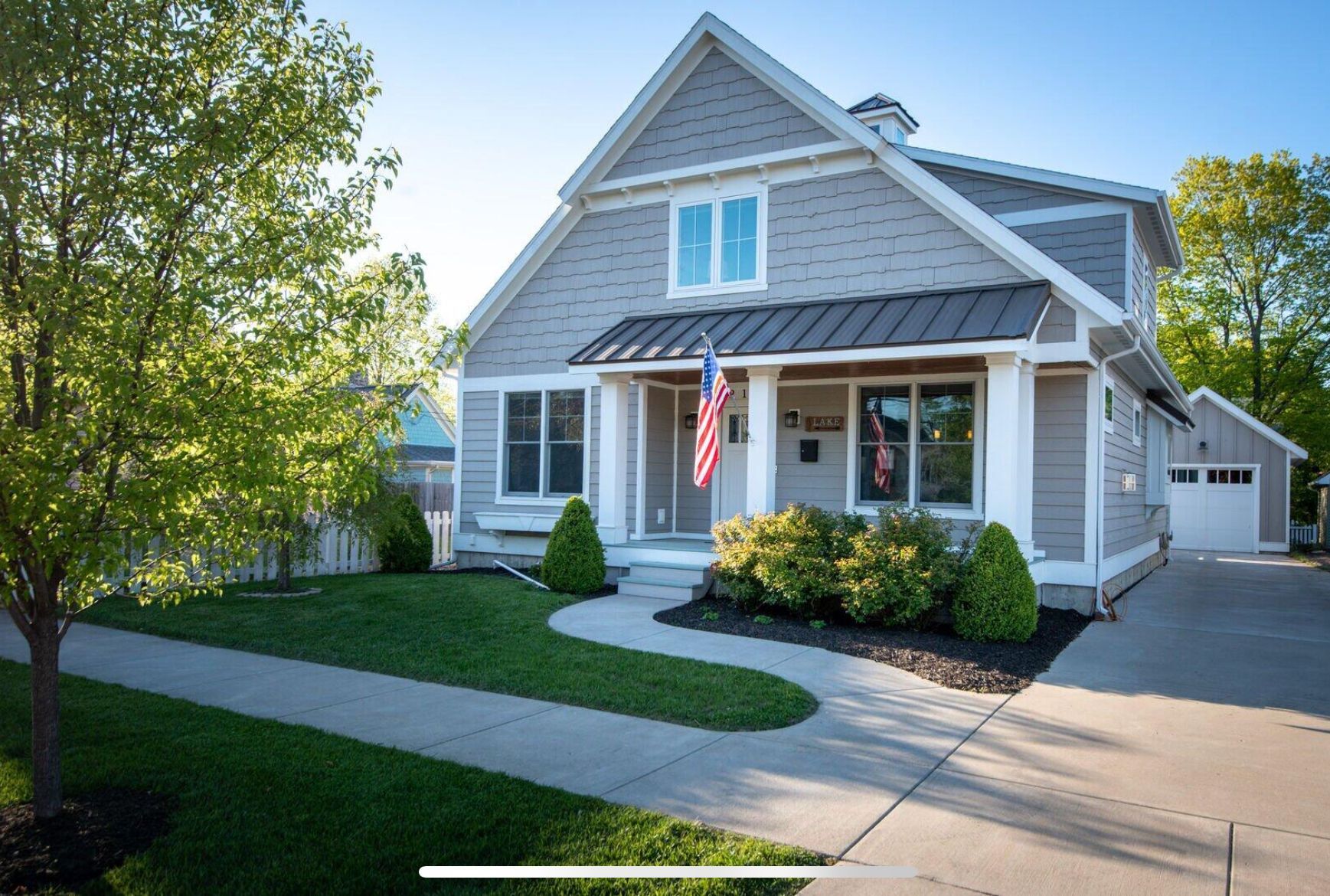 Gray suburban house with a front porch, American flag, and manicured lawn in bright daylight
