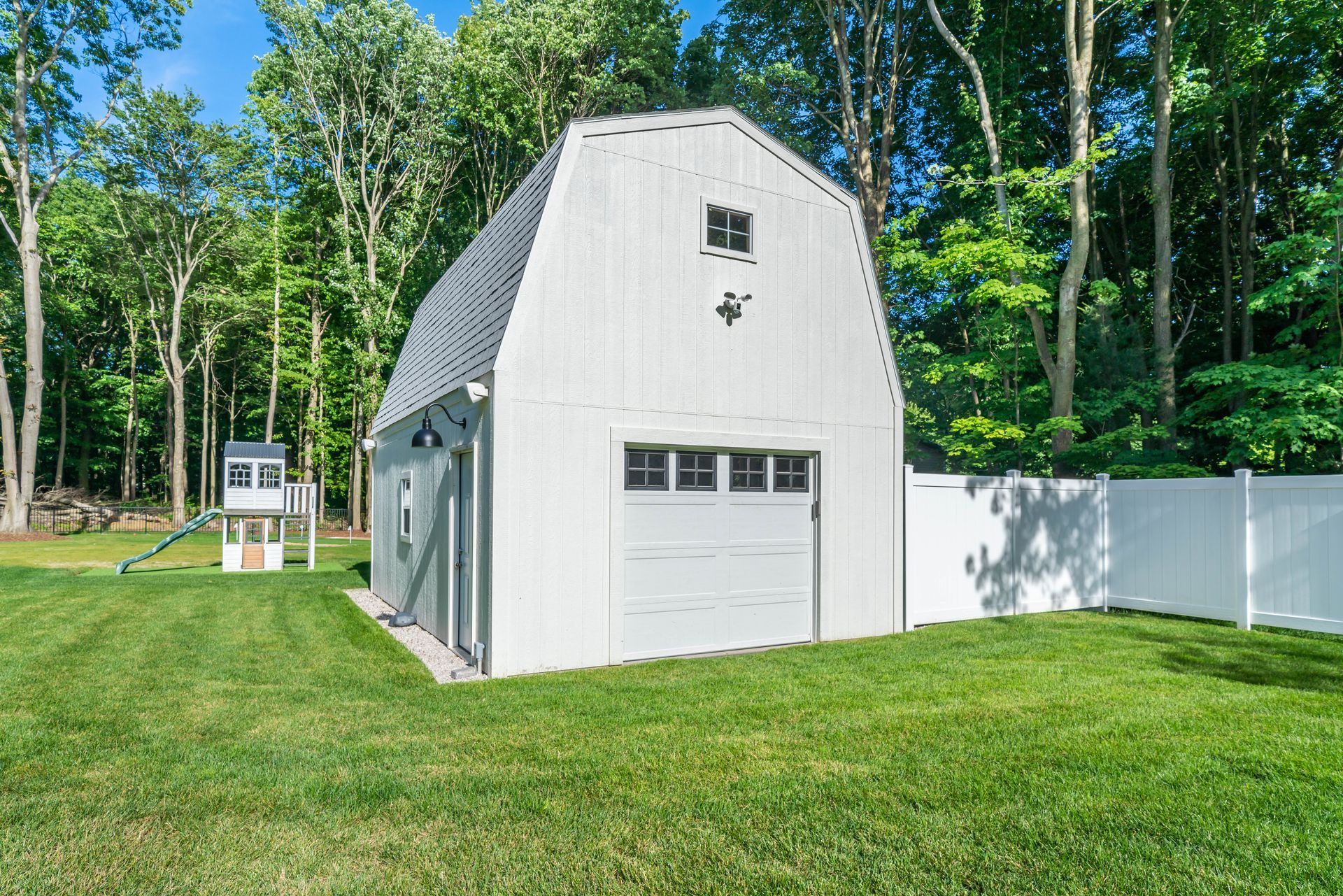 White barn-style shed with gray roof in a grassy backyard, surrounded by trees and a white fence