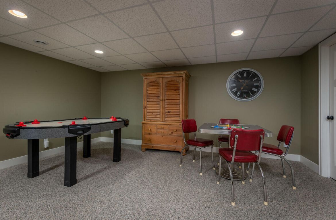 Basement rec room with air hockey table, wooden cabinet, and red chairs around a small table.