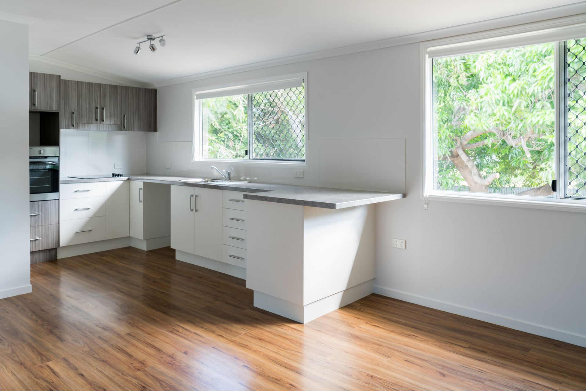 A modern kitchen with white cabinetry, wooden floors, and sunlight streaming through windows.