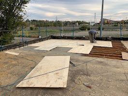 Construction worker laying plywood on an outdoor concrete surface with exposed wooden beams, surrounded by a fence and trees.