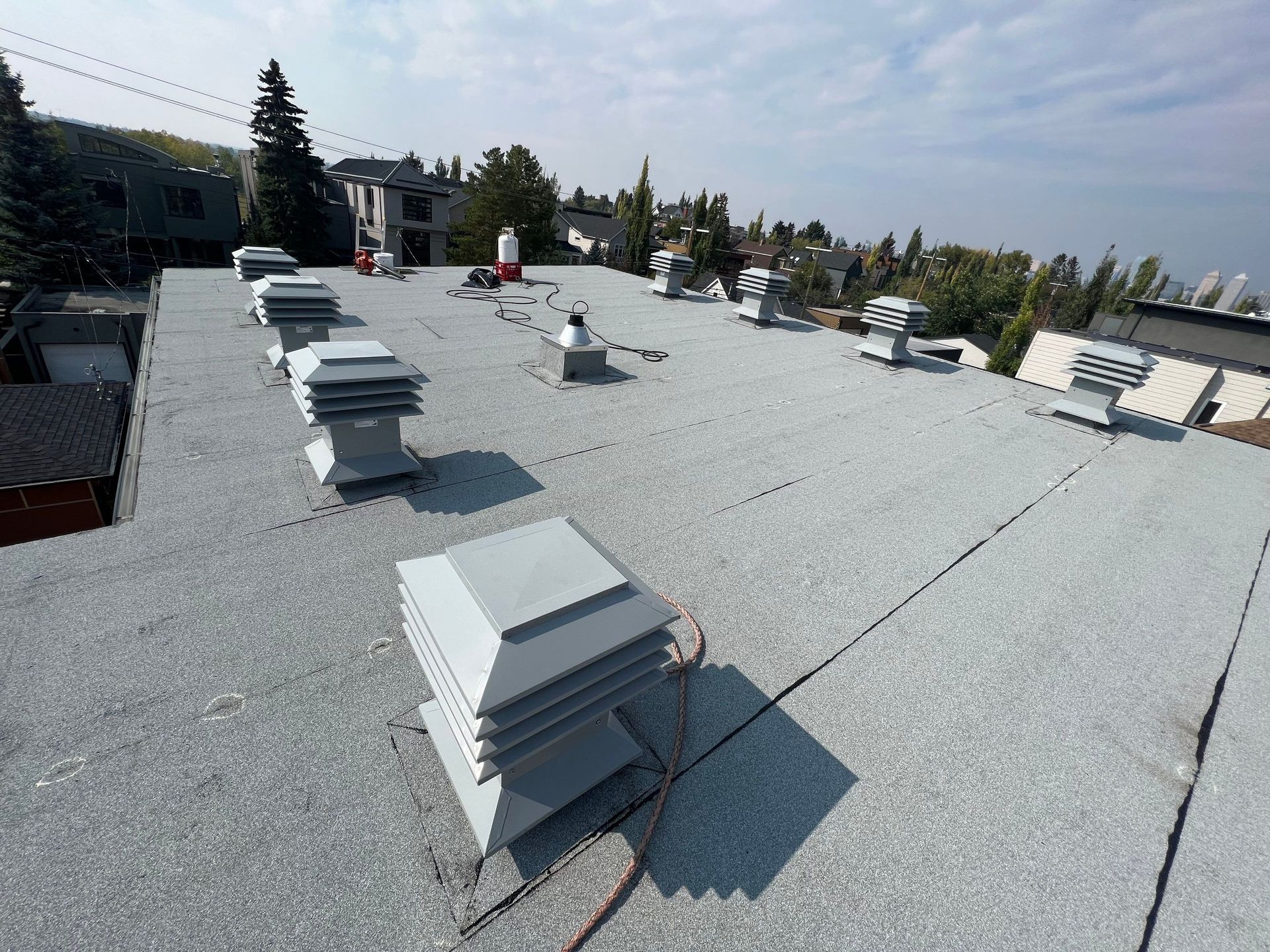 Gray rooftop with multiple square vents, trees, and buildings in the background under a clear sky.