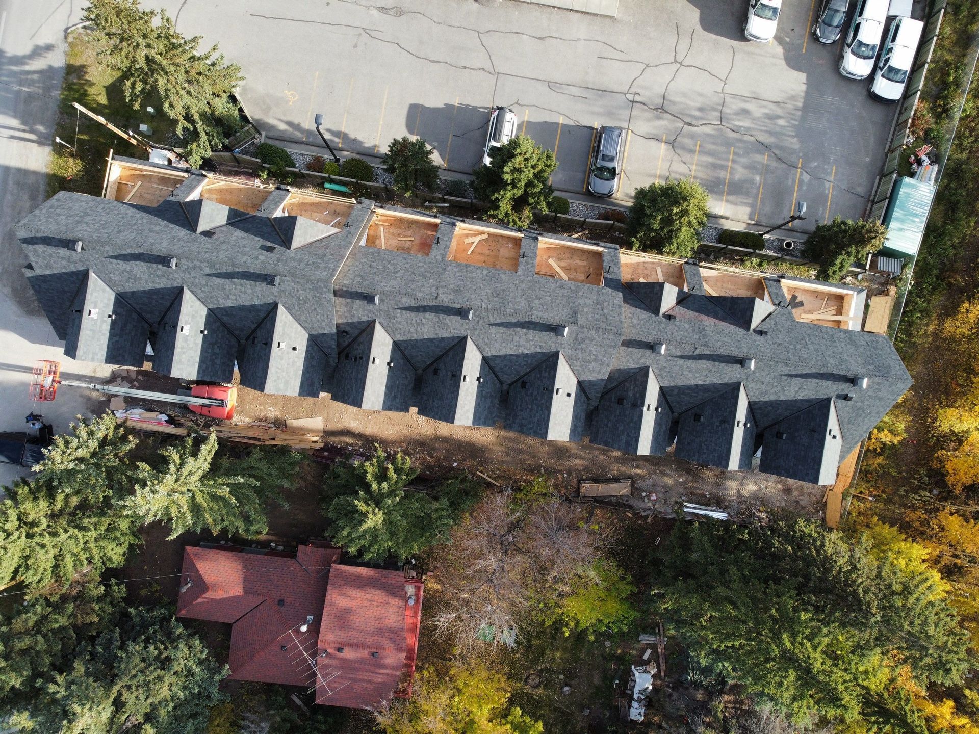 Aerial view of newly constructed townhomes with dark gray roofs, a red roofed house, and a parking area.