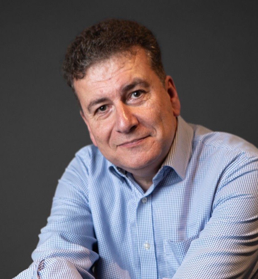Man in a light blue checkered shirt poses against a dark gray background, smiling gently.