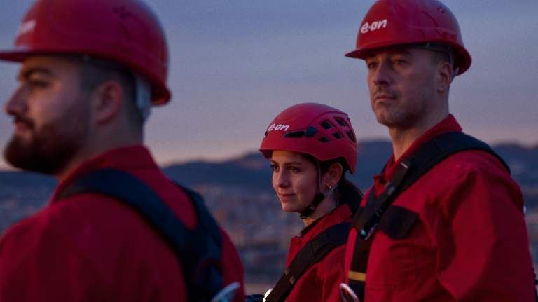 Three people in red helmets and jackets stand outdoors at dusk, looking serious.