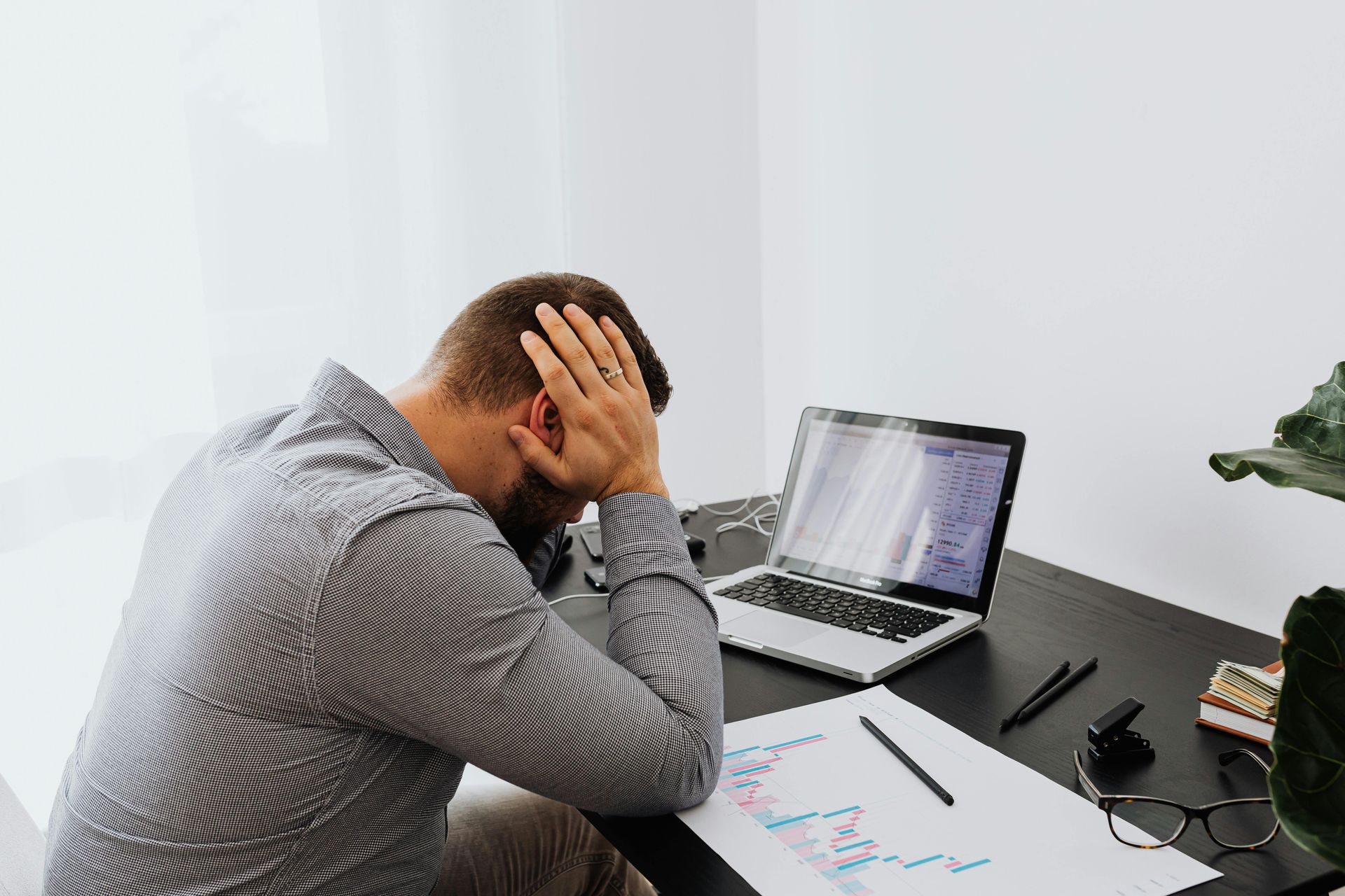 Stressed person at desk with laptop, head in hand, reviewing charts in a bright office