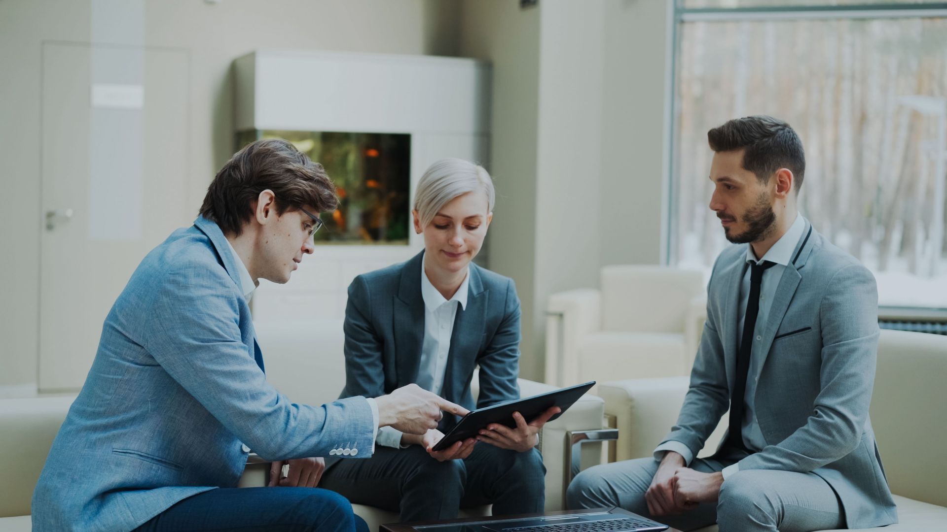Three coworkers in business attire discussing a tablet in a bright office lounge