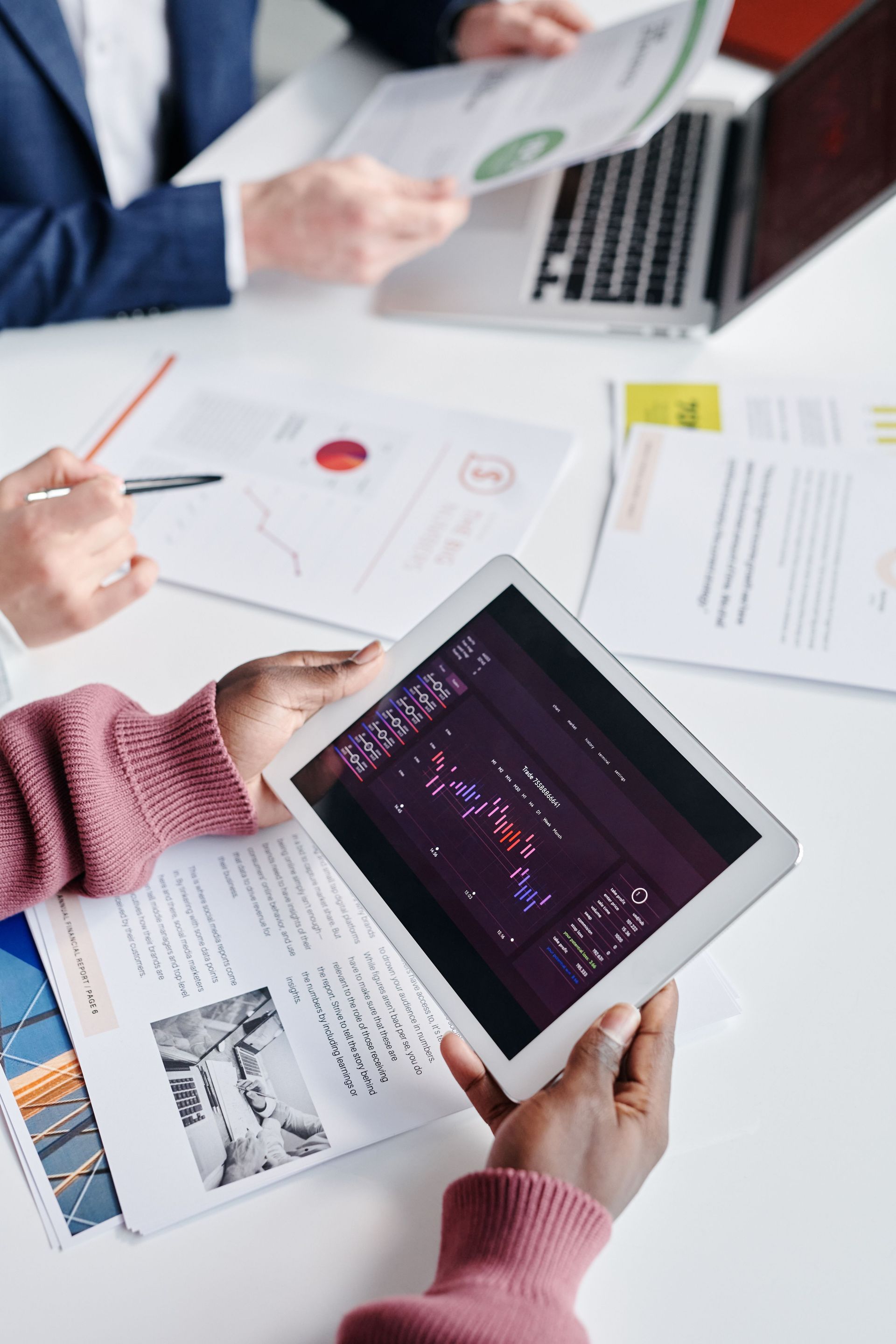 Hands holding a tablet with charts in a business meeting over papers and a laptop