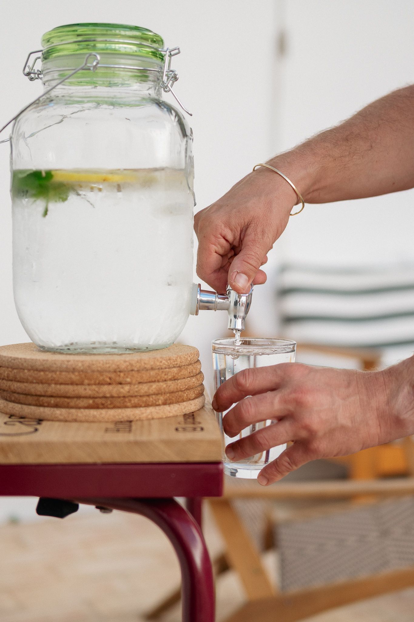 Person filling a glass with water from a clear beverage dispenser, set on a small red table.