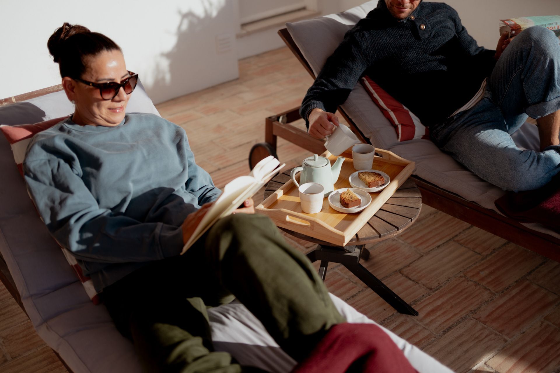Woman reading, man relaxing on patio; tea tray with pastries. Sunny outdoor setting.