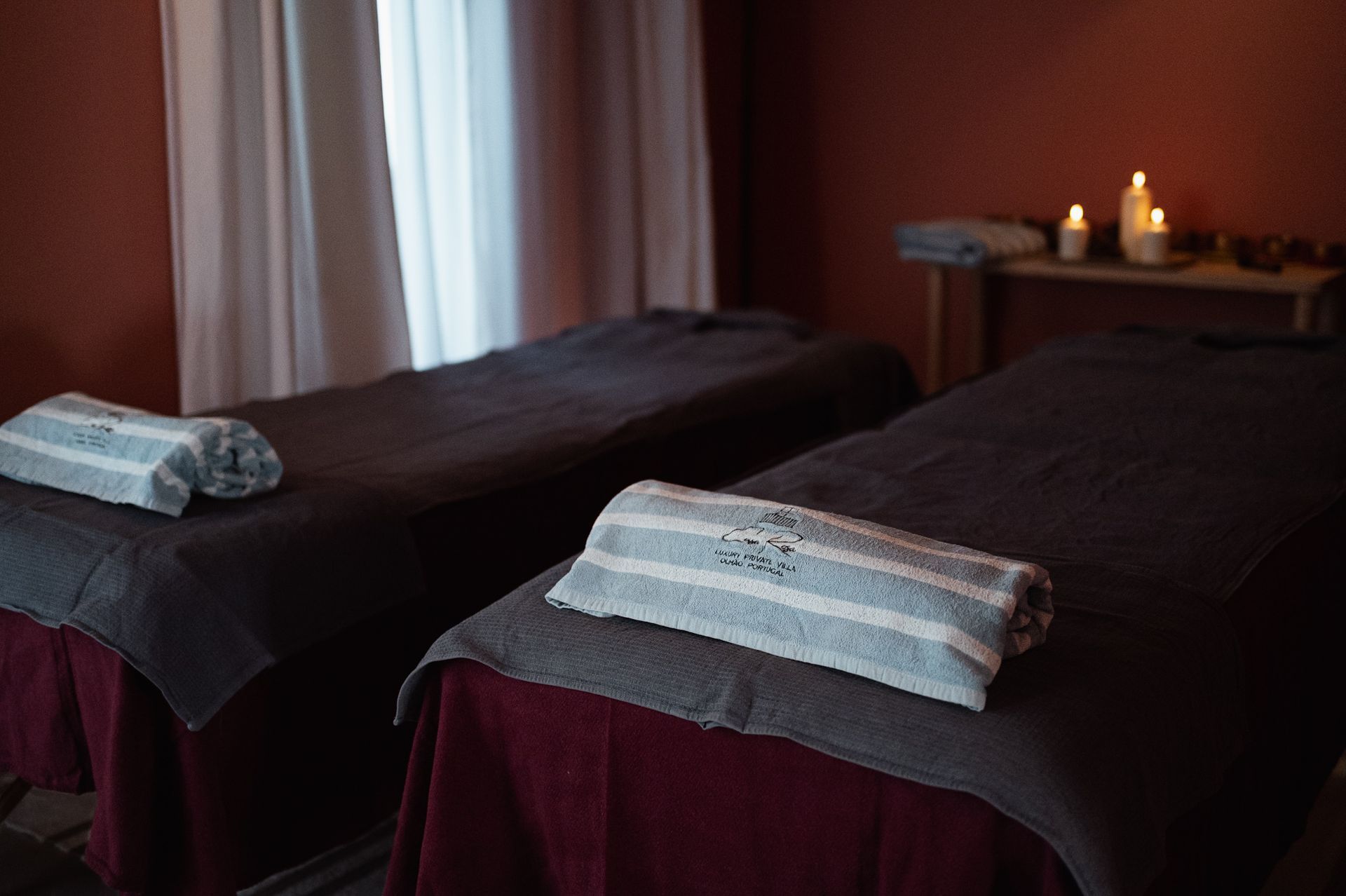 Two massage tables with towels in a dimly lit room, likely a spa. Candles and window present.