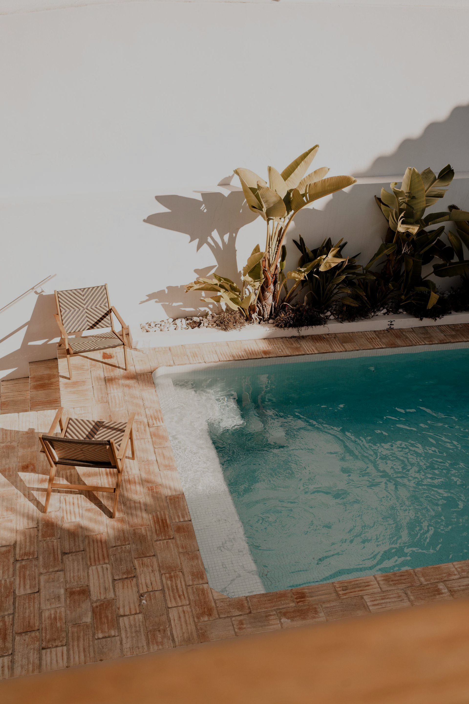 Pool with turquoise water, palm trees, and two chairs on a brick patio against a white wall.