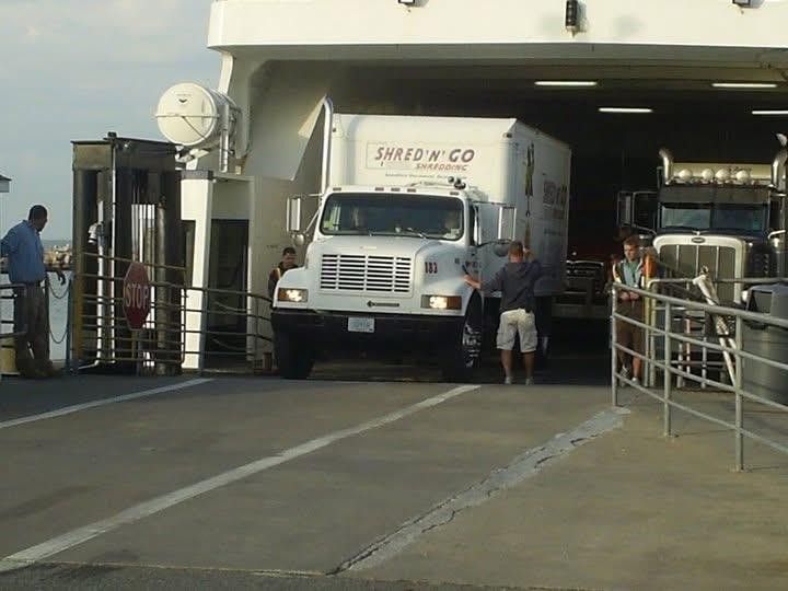 White truck driving off a ferry, with people nearby. Outdoors setting.