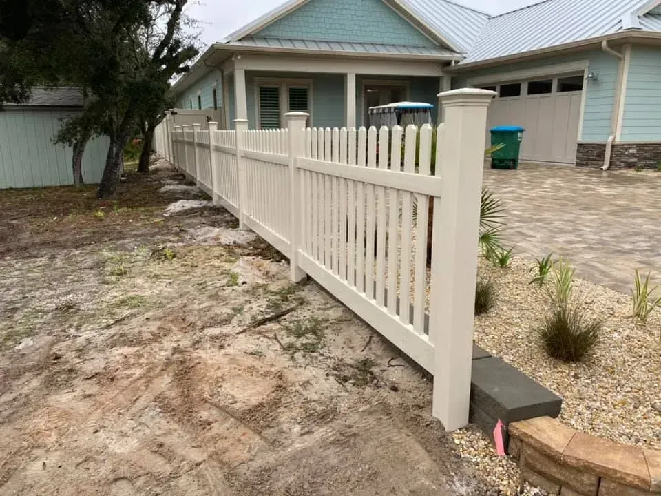 White picket fence beside a beige house with a blue roof on a sandy driveway