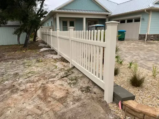 White picket fence bordering a driveway beside a light blue house with a front porch and sandy yard