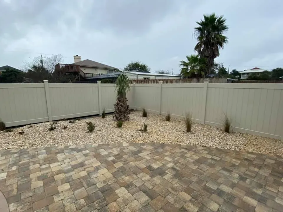 Paved backyard with beige fence, sparse young plants, and a palm tree under a cloudy sky