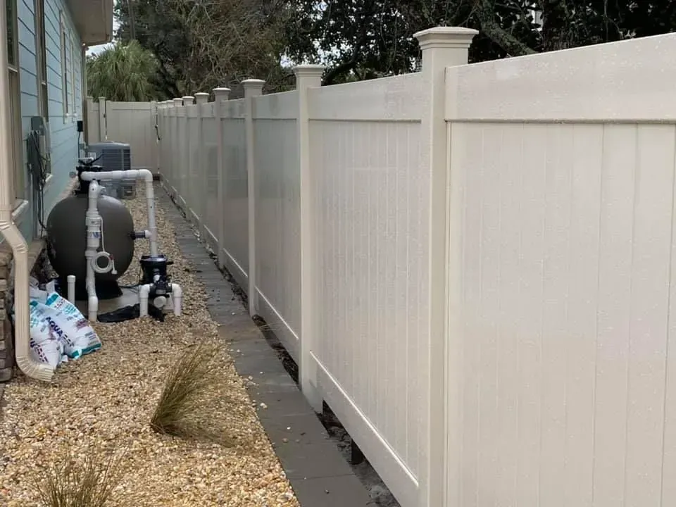 Narrow side yard with a white vinyl fence, gravel ground, and pool equipment beside a house