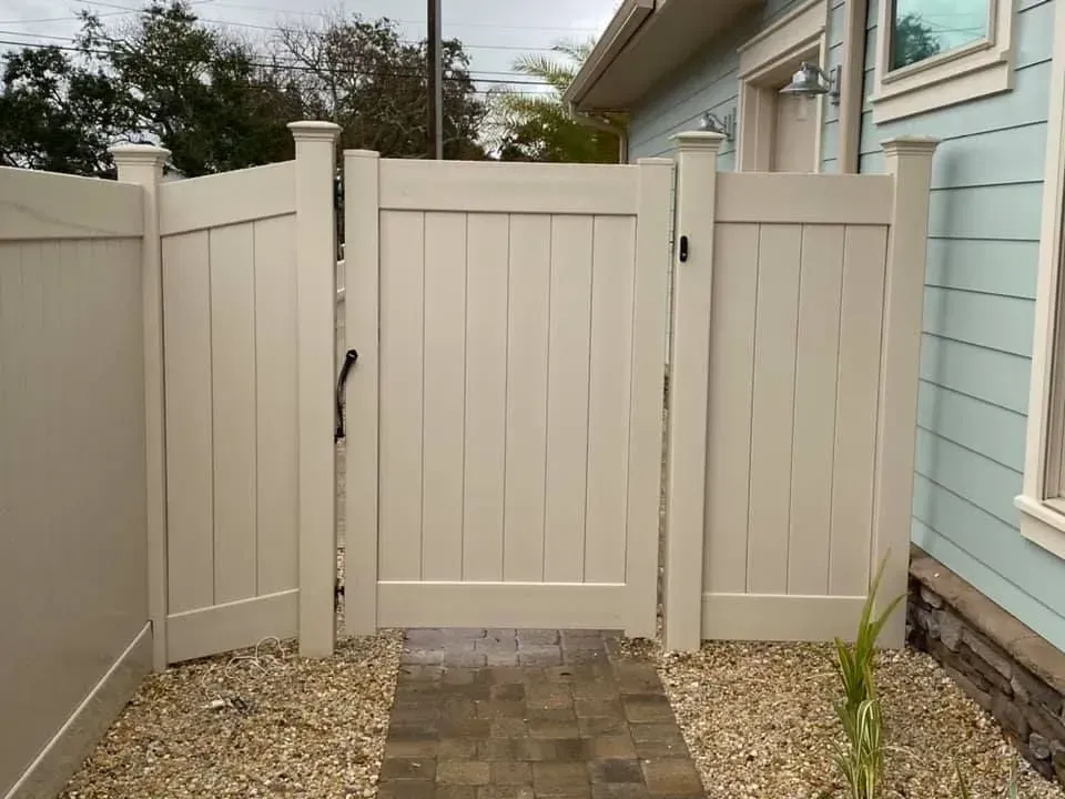 Closed beige backyard gate beside a blue house, with gravel and pavers in front