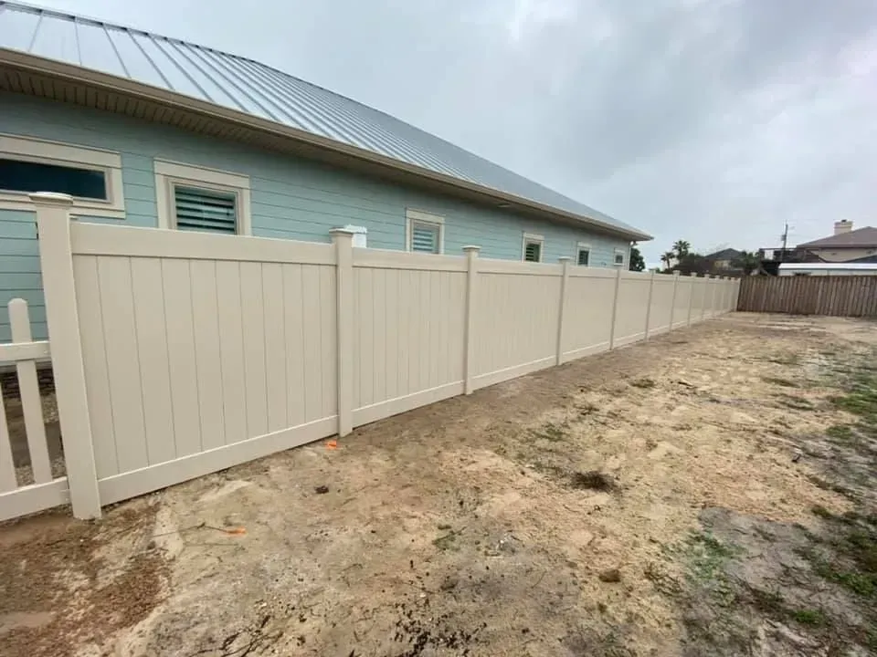 Beige privacy fence along a dirt side yard beside a light blue house under a cloudy sky