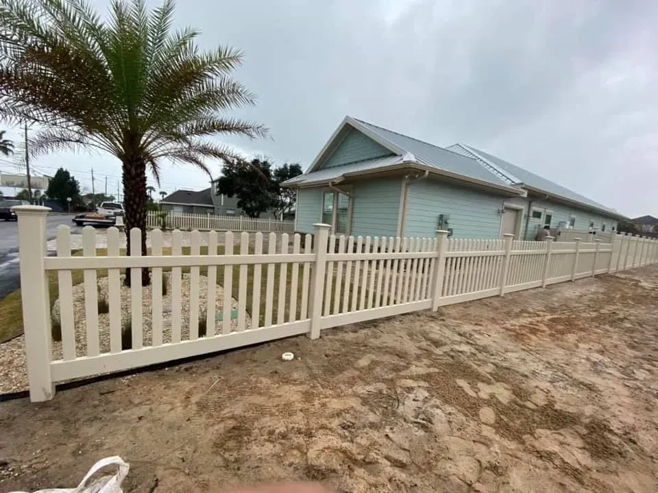 White picket fence beside a pale blue house with palm trees along a sandy road under cloudy skies