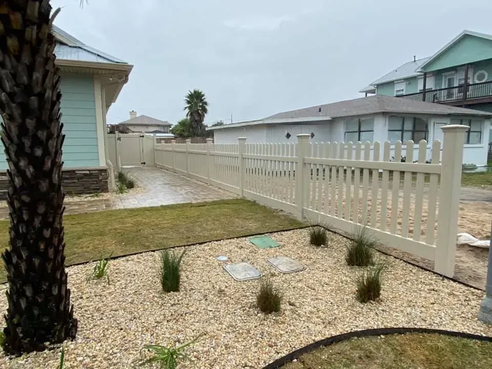 Small front yard with gravel, young plants, a white picket fence, and houses on a cloudy day.