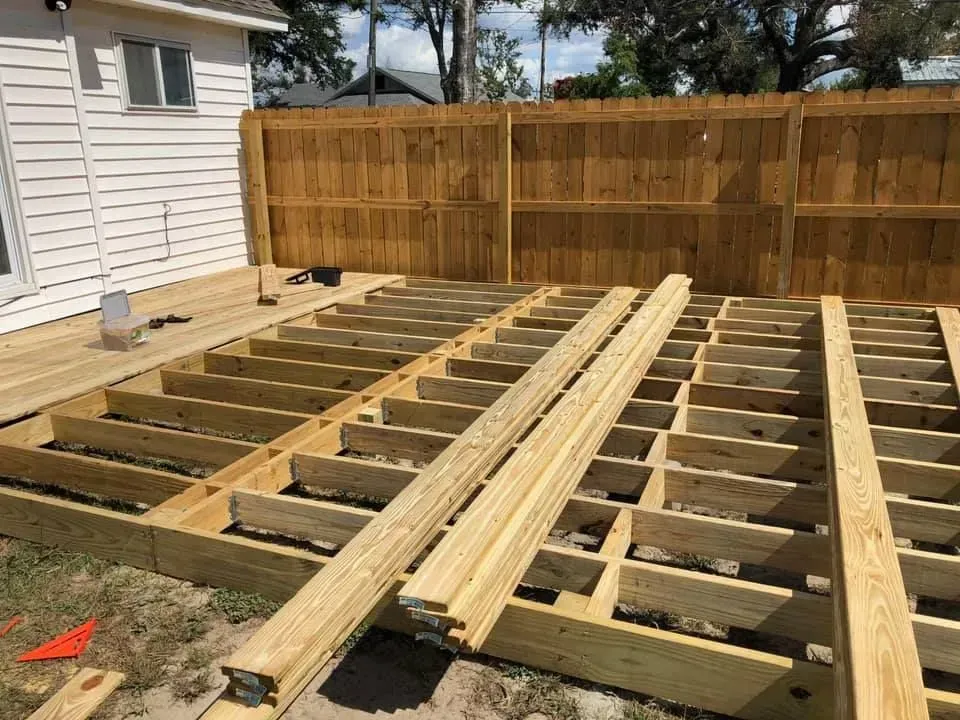 Wooden deck frame under construction beside a house, with lumber laid across the joists and a fenced backyard.
