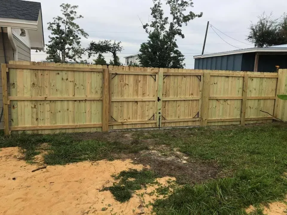 Wooden privacy fence with double gate in a grassy yard beside a house, under an overcast sky