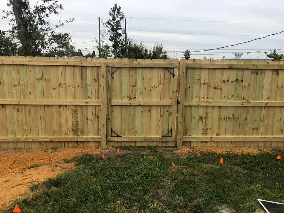 Wooden backyard fence with a centered gate, grass in front, and trees under a cloudy sky.
