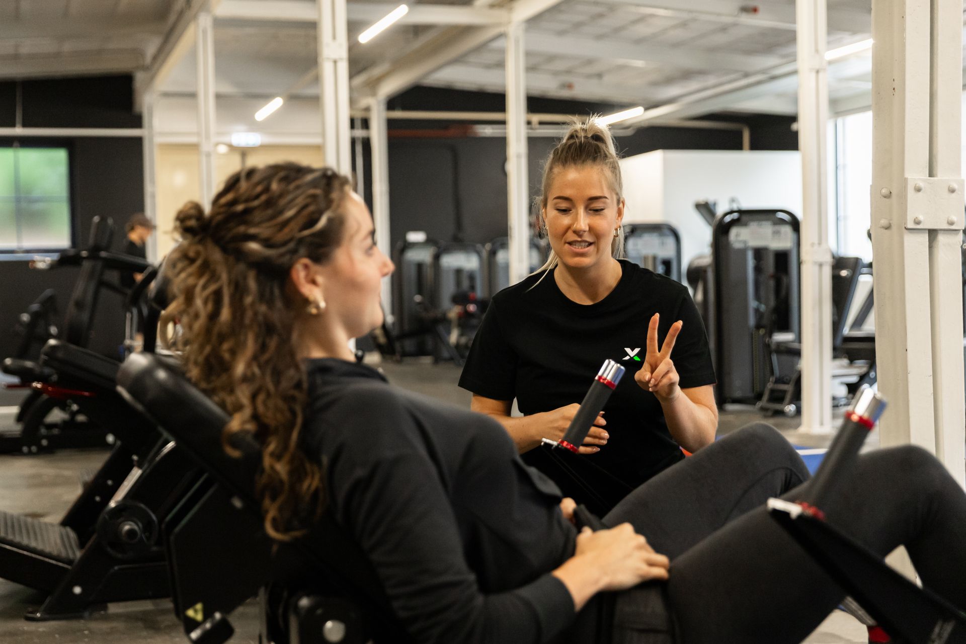 Two women talking in a gym, one seated on a machine and the other standing, holding a clipboard.