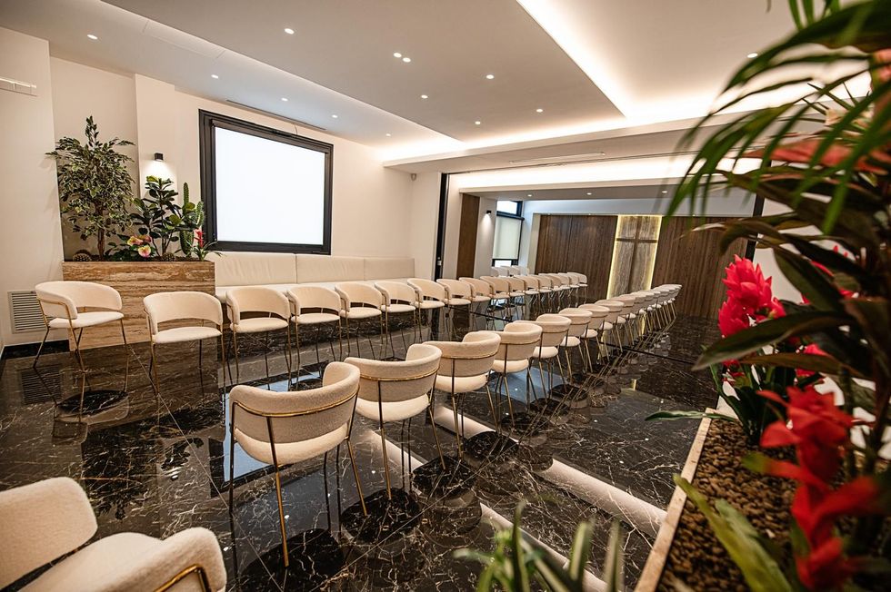 Sala conferenze moderna con file di sedie bianche, schermo per proiettore e piante lungo le pareti.