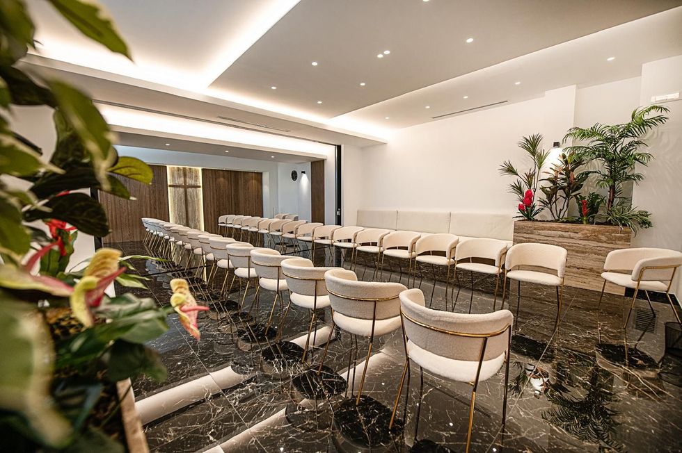 Sala conferenze moderna con file di sedie bianche, pavimento nero lucido e piante sotto faretti a incasso.